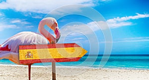 Spain flag on wooden table sign on beach background with pink flamingo. There is beach and clear water of sea and blue sky in the