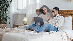 Spaniard arab married couple man and woman lying on bed in cozy bedroom home using laptop for video call making internet
