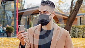 Spanish young man in medical mask talking on video call using smartphone standing in park hispanic guy nods approvingly