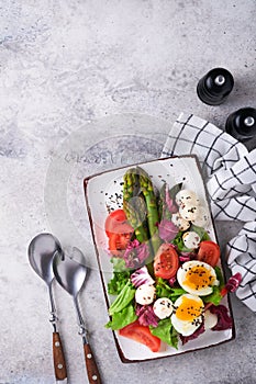 Sparagus, tomato, lettuce, mozzarella, black sesame, flax, oil olive salad and soft boiled egg on rectangular ceramic plate on