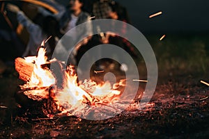 Sparking bonfire with tourist people sit around bright bonfire near camping tent in forest in summer night background. Group of