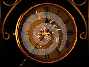 Sparkler in front of old golden vintage clock with roman numerals at 12 o'clock, New Year's Eve celebration