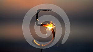 Sparkler in shape of number five 5 burning against backdrop of sea waves at dusk