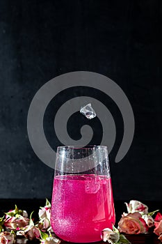 A sparkling brilliant raspberry drink in a glass stands on a wooden table with ice. In the background are roses and a garland