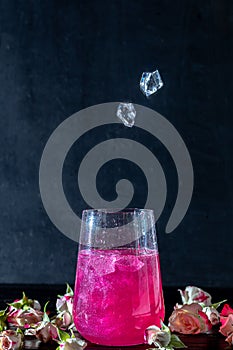 A sparkling brilliant raspberry drink in a glass stands on a wooden table with ice. In the background are roses and a garland