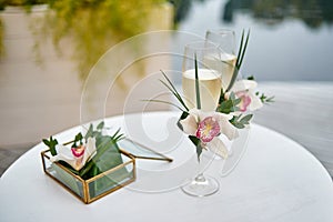 Sparkling wedding glasses with champagne, orchid flower decor and wedding rings in box in blur on white table outdoors, copy space