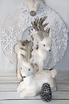 Sparkly fluffy white Christmas snow deer decorations in front of white wreath on white planks with silver pine cone - Selective