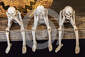 Speak no evil See no evil Hear no evil skeletons sitting on edge of a table with giant scarey metal pumpkin mouth behind them for