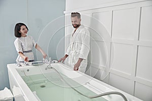 Specialist prepares hydro massage tub with clear water to mature guest in spa salon