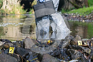 Specialist, in a protective suit and with a black suitcase, marks the crime scene numbers on the pond