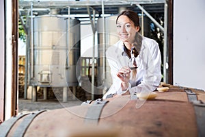 Specialist in white coat examines glass of wine on the background of barrels for fermentation