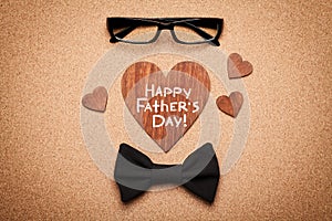 Spectacles, bowtie and wooden heart with note Happy Fathers Day, cork board background, top view, flat lay