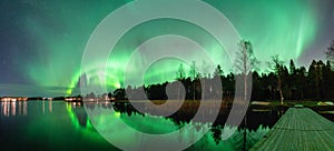 Spectacular night wide panorama strong green lights of dancing Aurora over Northern forest, reflection in lake, small boat, bridge