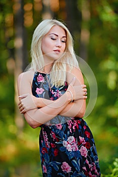 A spectacular young blonde woman with long hair elegantly poses outdoors against the background of trees in the summer.