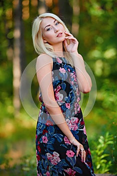 A spectacular young blonde woman with long hair elegantly poses outdoors against the background of trees in the summer.