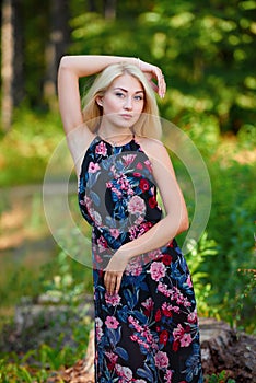 A spectacular young blonde woman with long hair elegantly poses outdoors against the background of trees in the summer.