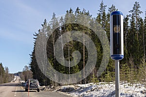 Speed camera on the roadside with a straight road and forest against blue sky in background