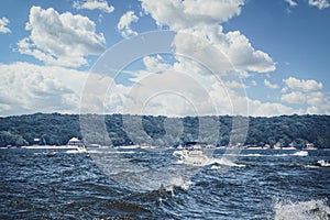 Speedboats and PWC churning up wakes on rough water at lake with wooded shore and lakeside homes in distance under blue sky with