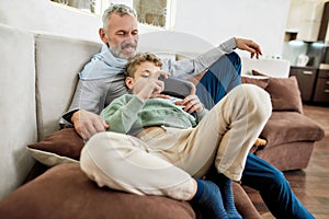 Spending time with grandson at home. Cute little boy playing video game on smartphone while relaxing on sofa with his