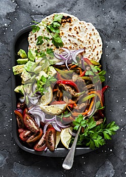 Spicy beef, vegetables, avocado, corn tortillas fajitas on a sheet pan on a dark background, top view. Delicious snack, tapas in