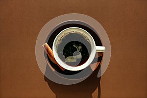 Spicy black coffee concept. Ceramic cup, saucer, cinnamon on chocolate brown background. Monochrome horizontal poster for cafe,