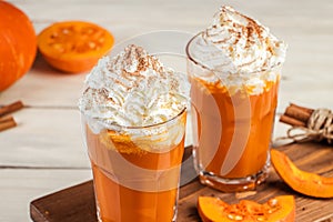 Spicy latte with pumpkin and whipped cream on wooden background. Hot coffee in glass mug and autumn leaves.