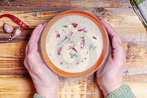 Spicy soup with sour cream, bread and spices on burned wooden table. Traditional rural dinner. Old woman hands holds clay bowl