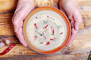 Spicy soup with sour cream, bread and spices on burned wooden table. Traditional rural dinner. Old woman hands holds clay bowl