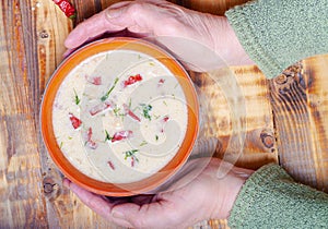 Spicy soup with sour cream, bread and spices on burned wooden table. Traditional rural dinner. Old woman hands holds clay bowl