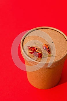 Spicy soup to go in a craft container with red pepper on a red background. Takeaway Food. Close up shot