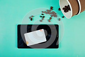 Spilled Coffee Beans Near a Takeaway Cup and Tablet With Credit Card on Teal Background