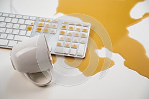 Spilled cup of black coffee on computer keyboard on white table.