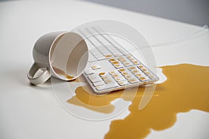 Spilled cup of black coffee on computer keyboard on white table.