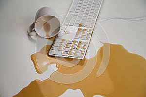 Spilled cup of black coffee on computer keyboard on white table.
