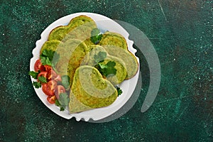 Spinach avocado pancakes in the shape of a heart with greek yogurt sauce and cherry tomatoes on a black plate over dark background