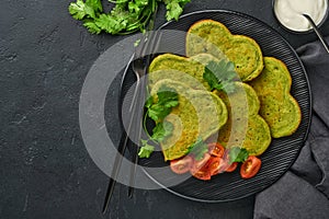 Spinach avocado pancakes in the shape of a heart with greek yogurt sauce and cherry tomatoes on a black plate over dark background