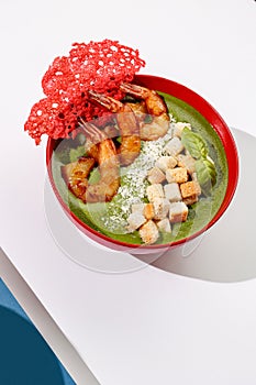 Spinach cream soup with shrimps, parmesan and crackers in red color bowl on white background with sunlight and shadows, minimalism