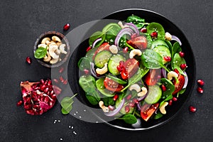 Spinach salad with fresh cucumbers, tomato, onion, pomegranate, sesame seeds and cashew nuts on black background. Healthy vegan fo