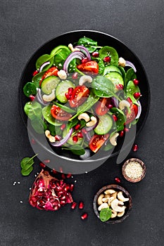 Spinach salad with fresh cucumbers, tomato, onion, pomegranate, sesame seeds and cashew nuts on black background. Healthy vegan fo