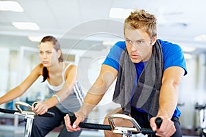Spinning exercise. Young man training on exercise bike with woman in background.