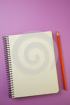 Spiral-bound notebook and orange pencil lie on a lilac background