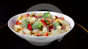 Spiral pasta with vegetables on a plate revolve on a black background. Green cauliflower falls on a plate