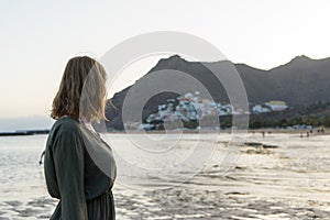 Spiritual adolescent in green dress admire the sunset and the sea town on a hill in Tenerife. Blonde romantic woman look the