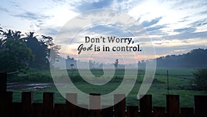Spiritual inspirational quote - Don\'t worry, God is in control. On beautiful background of the sunrise over the green field.