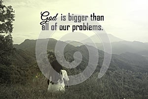 Spiritual inspirational quote - God is bigger than all of our problems. With person sitting alone from behind in the mountains.