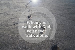 Spiritual inspirational quote - When you walk with God, you never walk alone. With footprints on beach black sand background.