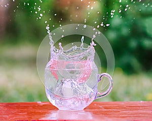 Splash of cool fresh water with ice cube in transparent glass cup in the table outoors in summer day.