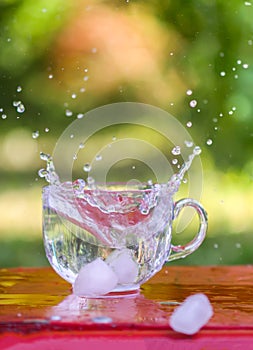 Splash of cool fresh water with ice cube in transparent glass cup in the table outoors in summer day.