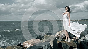 Splashes of water envelops an attractive young woman. Beautiful girl in a white dress stands on seashore