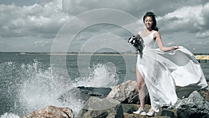 Splashes of water envelops an attractive young woman. Beautiful girl in a white dress stands on seashore